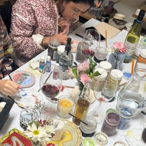Mesa com tintas, flores, taças de vinho e pratos sendo pintados